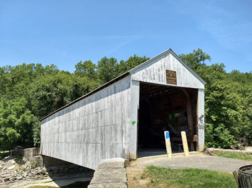 A wooden covered bridge with a sign, surrounded by green trees and a clear blue sky.