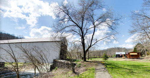A scenic view of a white barn by a stream, with a tree and a path leading to a red and orange building in the background.