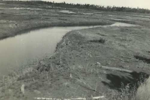 A winding river flows through a barren landscape, with sparse vegetation and distant trees under a clear sky.