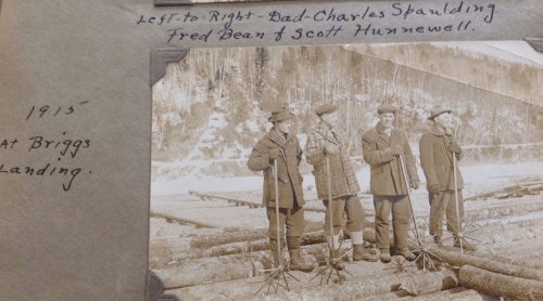 Four men standing on logs at a landing in 1915, holding tools, with handwritten notes above and below the photo.