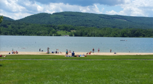 A sunny beach scene with people swimming and relaxing by a lake, surrounded by green hills and blue skies.