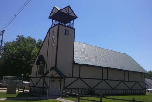 A large, tan building with a green roof and a bell tower, surrounded by grass and a wooden fence.