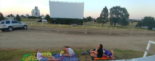 A group of friends relaxes on blankets at an outdoor movie theater, with a large screen in the background.