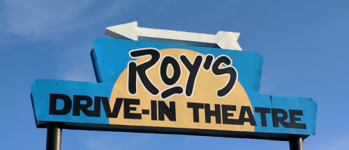 Sign for Roy's Drive-In Theatre with a blue background and an arrow pointing left against a clear sky.