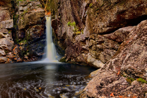 A serene waterfall cascades into a dark pool, surrounded by rugged rocks and autumn foliage.