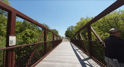 A wooden bridge surrounded by greenery on a sunny day, with a person walking on the right side.
