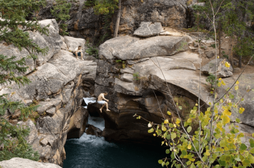 Two people relax on rocky cliffs above a flowing river, surrounded by trees and autumn foliage.