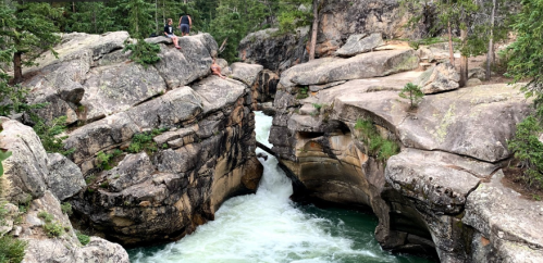 Two people sit on rocky ledges overlooking a rushing river flowing through a narrow canyon surrounded by trees.