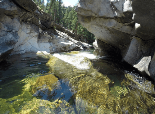 A serene river flows between rocky cliffs, surrounded by lush green trees under a clear blue sky.