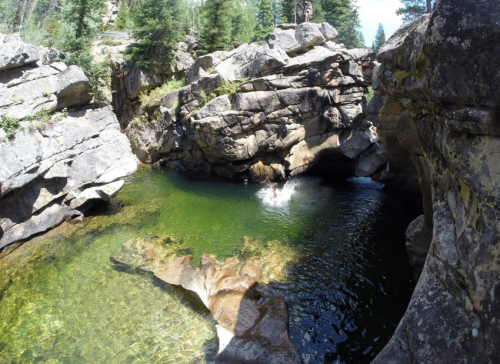 A person jumps into a clear, green pool surrounded by large rocks and trees in a natural setting.