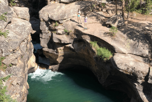 Two people stand on a rocky ledge above a deep, turquoise pool surrounded by cliffs and trees.