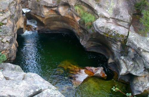 A serene natural pool surrounded by rocky cliffs and lush greenery, with water flowing gently over the rocks.