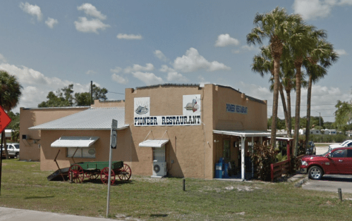 A small, tan building with a sign reading "Pioneer Restaurant," surrounded by palm trees and parked cars.