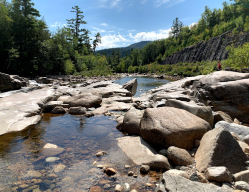 A serene river scene with smooth rocks, clear water, and lush greenery under a blue sky with scattered clouds.