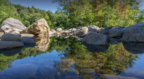 A serene landscape featuring a calm pool of water surrounded by large rocks and lush green trees reflecting in the water.