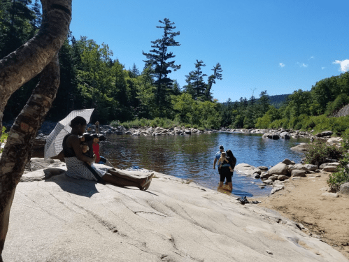 A sunny riverside scene with people enjoying the water, surrounded by trees and rocky terrain. One person relaxes on the shore.
