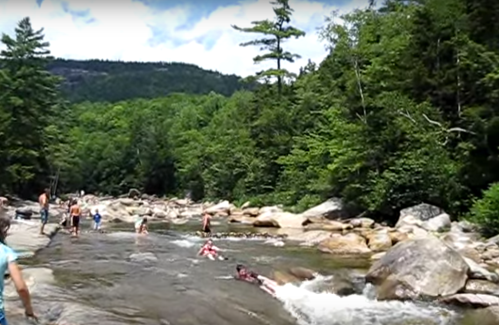 People swimming and playing in a rocky river surrounded by lush green trees and mountains under a blue sky.