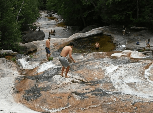 A scenic view of a rocky river with people swimming and climbing, surrounded by lush green trees and mountains.
