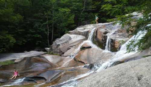 A rocky waterfall with two people exploring the area, surrounded by lush green trees.