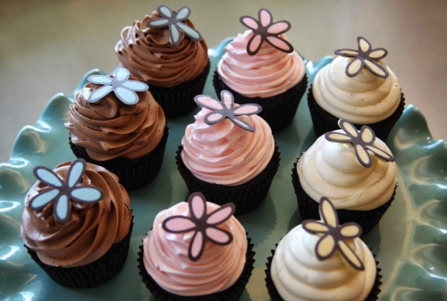 A plate of cupcakes with chocolate, pink, and vanilla frosting, each topped with a flower-shaped decoration.
