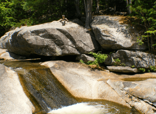 A person sits on a large rock by a flowing stream surrounded by trees and greenery.
