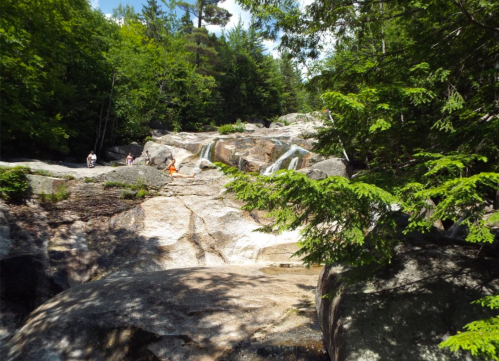 A scenic view of a rocky waterfall surrounded by lush green trees and people enjoying the natural landscape.