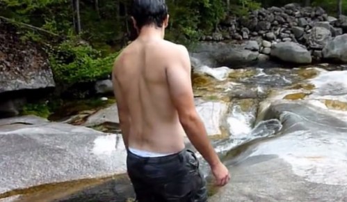 A person with a bare back stands near a rocky stream, surrounded by trees and smooth stone surfaces.