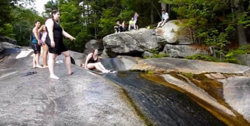 A group of people enjoying a natural water slide on smooth rocks, surrounded by trees and a rocky landscape.