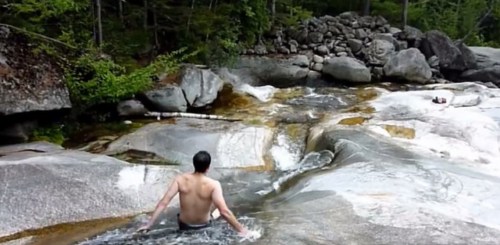 A person sits in a natural water pool surrounded by smooth rocks and greenery, enjoying a sunny day outdoors.