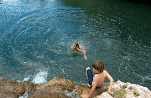 A child sits on a rock by the water while another swims nearby in a natural setting.