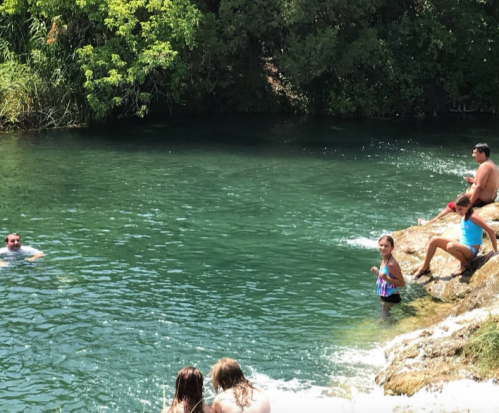 People enjoying a sunny day by a clear, greenish-blue swimming area surrounded by lush greenery.