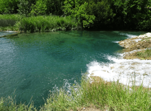 A serene river with clear turquoise water, surrounded by lush greenery and rocky banks. Gentle waves flow over the rocks.