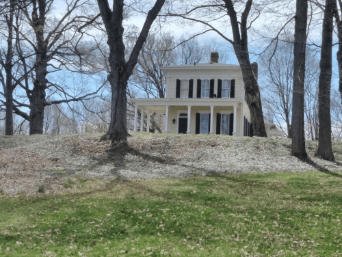 A two-story yellow house with black shutters, surrounded by trees and a grassy area under a clear blue sky.