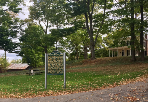 A historical marker stands in a park with trees and a house in the background, surrounded by fallen leaves.
