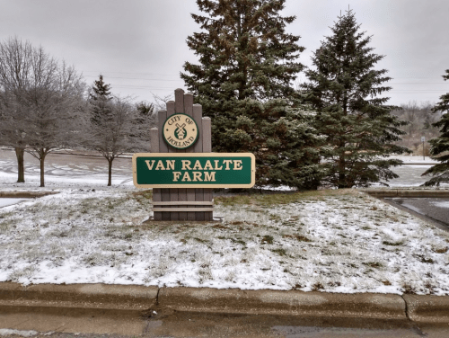 Sign for Van Raalte Farm in the City of Holland, surrounded by snow and evergreen trees.