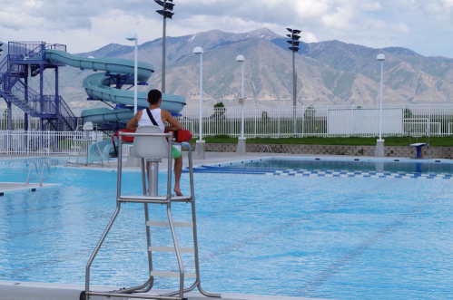 A lifeguard sits on a tall chair by a pool with a water slide, overlooking the water and mountains in the background.