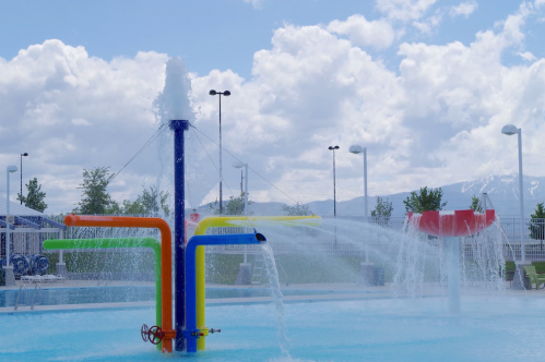 Colorful water fountain with spray features in a pool area, under a bright sky with fluffy clouds.