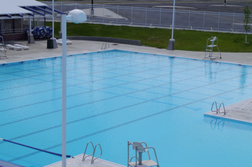 A clear, empty swimming pool with lane markings, diving boards, and poolside chairs under a sunny sky.