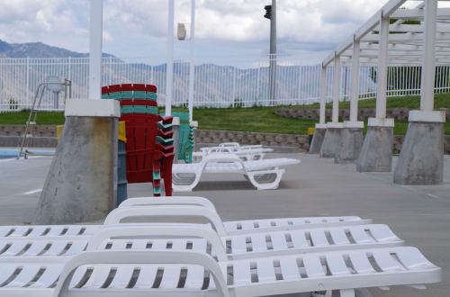 Empty poolside area with white lounge chairs and colorful stacked chairs against a backdrop of mountains and cloudy sky.