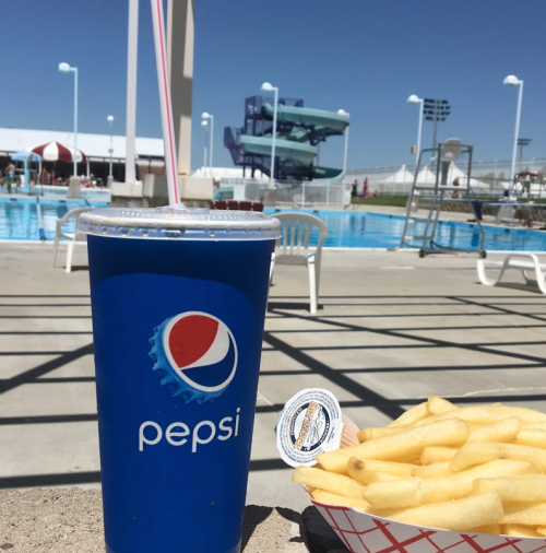 A large Pepsi cup and a tray of fries sit on a poolside table, with a water slide and pool in the background.