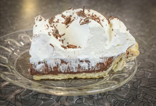 A slice of chocolate cream pie topped with whipped cream and chocolate shavings on a clear glass plate.