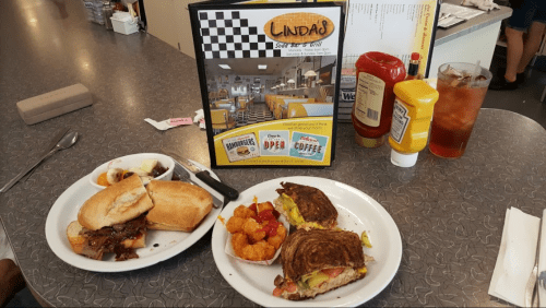A table with two plates of food: a sandwich, tater tots, and a side dish, alongside a menu and condiments.
