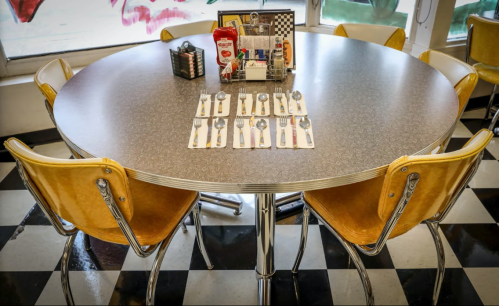 A round diner table set with spoons and napkins, surrounded by yellow chairs in a retro-style setting.