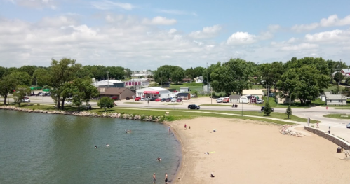 A sandy beach by a lake with people swimming, surrounded by green trees and buildings in the background under a blue sky.
