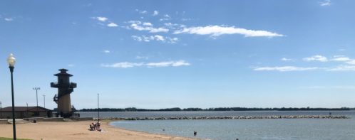 A sandy beach with people relaxing, a lighthouse tower in the background, and a calm lake under a clear blue sky.