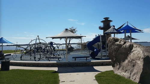 A playground by the water featuring climbing structures, slides, and shaded areas under blue skies.