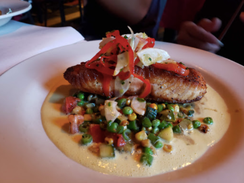 A plated dish featuring a seared fish fillet on a creamy sauce, topped with vegetables and garnished with red peppers.