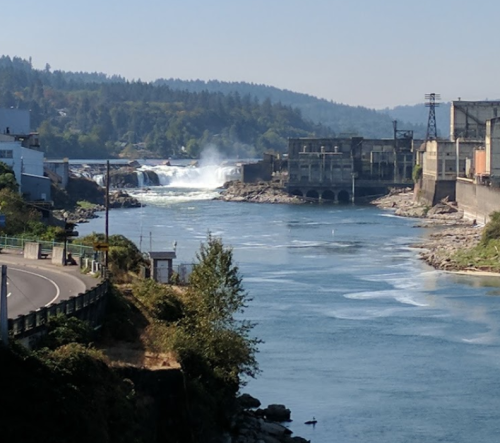 A river flows past a dam and waterfall, surrounded by trees and industrial buildings under a clear blue sky.
