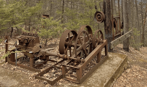 Rusty vintage machinery sits in a forest, surrounded by trees and fallen leaves, showcasing industrial relics of the past.