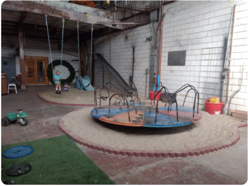 Indoor playground featuring a merry-go-round, swings, and a slide, with sand and play equipment in a spacious area.